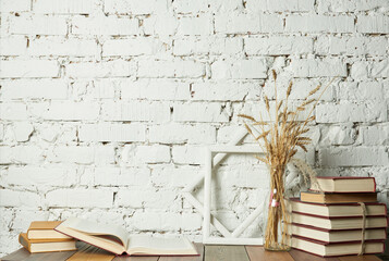 Wooden shelf with stack of books, wooden frames and dry spikelets in glass vase against white brick wall background