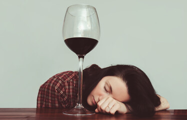 A young caucasian girl in a plaid shirt with tousled hair sleeps lying on a wooden table about a large glass of red wine. Concept of alcohol abuse, alcoholism, hangover, loneliness and depression.