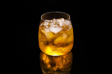 Glass with yellow cocktail with ice on black reflective surface