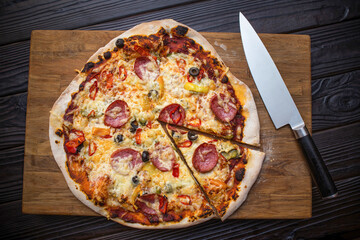 Homemade pizza and a large knife on an oak board and on a black textured table. An appetizing slice has been cut from the pizza.