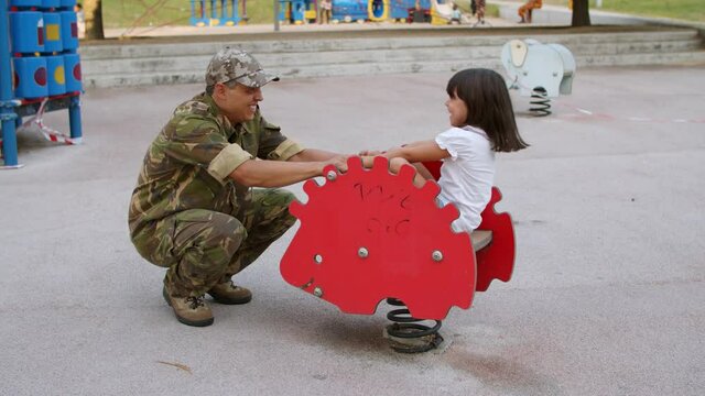 Military daddy playing with active kid outdoors. Girl enjoying time with her dad, riding spring rocking hedgehog on playground. Parenthood or childhood concept