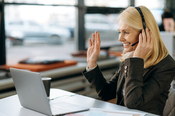 Middle aged female employee sit at the work desk, having a video conference with colleagues, waves hand and greeting. Friendly charming woman wearing headset, works as employee of support line, smiles