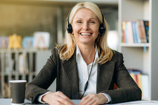 Webcam portrait of successful joyful female consultant in headset at the workplace. Happy senior adult business lady in stylish suit sit at the desk, communicate with colleagues on video conference