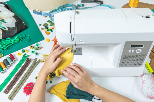 Top View Of Hands Sewing Fabric Masks On Sewing Machine. Dressmaker's Workspace. Make Your Own Face Mask At Home. Elastic, Threads, Textile, Buttons And Measuring Tape.