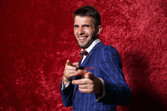 Handsome male master of ceremonies in elegant blue suit pointing at camera while acting during show on red background 
