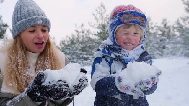 Beautiful cheerful mommy and son holding snow in hands and blowing it out on camera, making little snowstorm, having fun, enjoying pastime in snowy forest, winter moments, slow motion