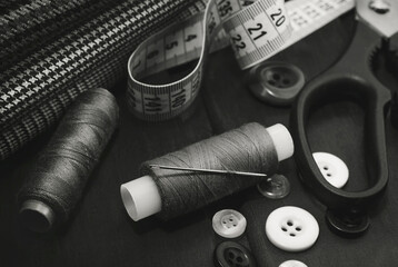 A needle with thread, buttons and scissors lie on a wooden background. Black and white photo. The working tools of a tailor or a seamstress.