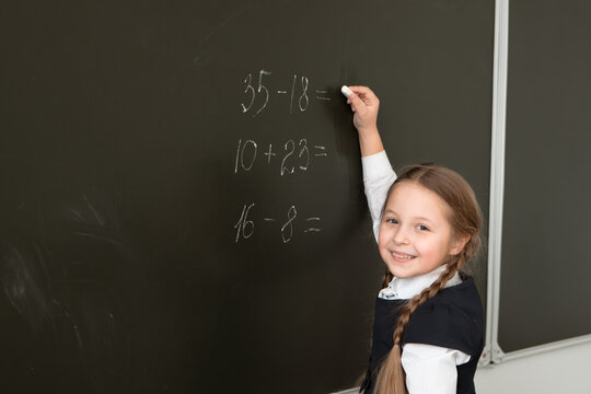 smiling little girl solve math task in front of blackboard in classroom of primary school 