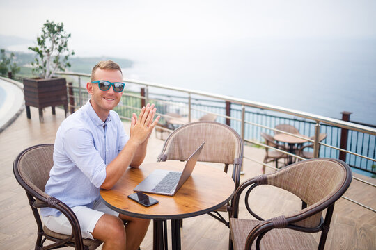 Handsome successful young male businessman sitting at a table by the pool with a laptop overlooking the Mediterranean Sea. Remote work on vacation. Vacation concept