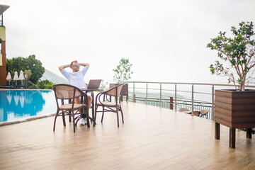 Handsome successful young male businessman sitting at a table by the pool with a laptop overlooking the Mediterranean Sea. Remote work on vacation. Vacation concept