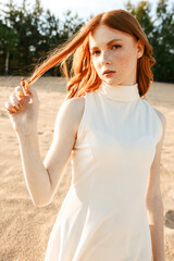 Delicate female model in stylish summer dress and with freckles standing on sand dunes in nature and touching long red hair while looking at camera 
