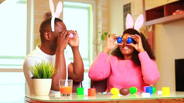 Young african american man and woman wearing cute easter bunny ears and painting eggs in the house