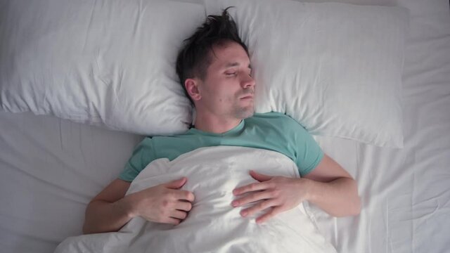 The young man does not sleep well. Nightmares. A bad dream. Worry. White bedding, turquoise blue t-shirt. Dark hair. Top view