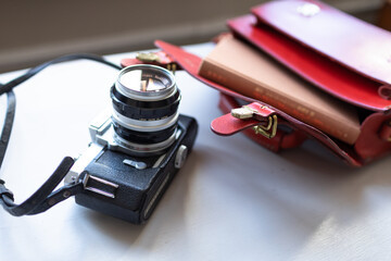 Antique film camera, book and red leather bag on a white table by the window. Stylish hobby time.