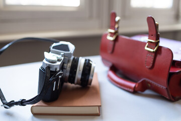 Antique film camera, book and red leather bag on a white table by the window. Stylish hobby time.