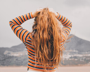 chica joven y bonita vestida con una blusa naranja se recoge el cabello de espaldas con un par de montañas detras