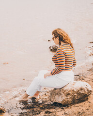 chica joven y bonita  vestida con una blusa naranja y un pantalón blanco sentada en una roca en una pequeña playa con un lago en frente