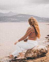 chica joven y bonita  vestida con una blusa naranja y un pantalón blanco sentada en una roca en una pequeña playa con un lago en frente