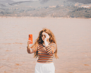 Chica joven y bonita vestida con una blusa naranja y un pantalón blanco tomándose una fotografía de viaje en una playa con un lago atrás
