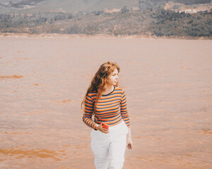 Chica joven saliendo del agua agarrando su celular vestida con un pantalón blanco  y una blusa naranja.
