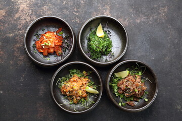 Composition of appetizers: tuna tartare, salmon tartare, kimchi, seaweed Japanese salad on a black stony background.