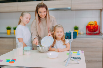 Obraz na płótnie Canvas Mom teaches her daughters to cook dough in the kitchen. The family is filming a culinary video on a smartphone.