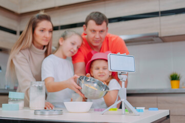 Mom with Father teaches two daughters to cook dough in the kitchen. The family is filming a culinary video on a smartphone.