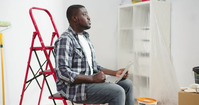 Side view of concentrated African American young handsome man tapping browsing on laptop computer surfing internet sitting on red ladder, house renovation, repair concept, portrait, decorating room