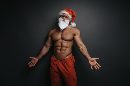 Shirtless muscular man in Santa hat with holding his hands to side. Sexy Christmas hunk in studio at grey background.