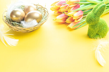 Golden eggs in basket with spring tulips, white feathers on pastel yellow background in Happy Easter decoration. Foil minimalist egg design, modern design.