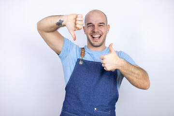 Young bald man wearing apron uniform over isolated white background Doing thumbs up and down, disagreement and agreement expression. Crazy conflict