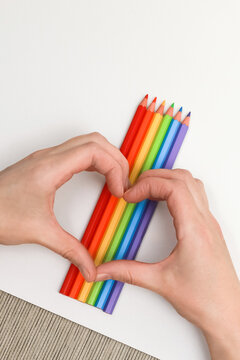 Female hands show the shape of a heart on a background of a rainbow made of colored pencils and a white sheet of paper diagonally, close-up, copy space