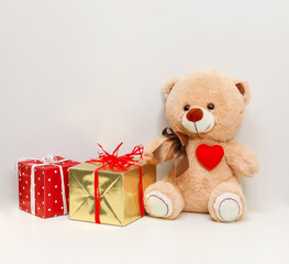 Plush Teddy bear with a soft fabric heart and a bow surrounded by two boxes with gifts in gold and red polka dot packaging, on a light background, copy space