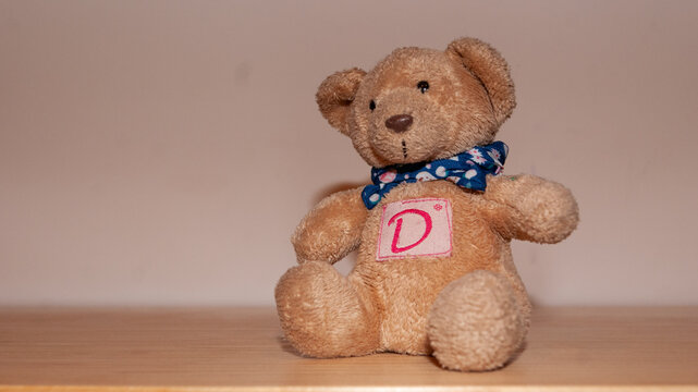 Osito de peluche con una letra "D" en el pecho sentado sobre mesa de madera