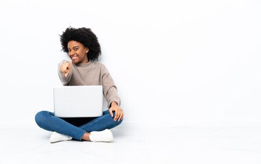 Young African American woman with a laptop sitting on the floor points finger at you with a confident expression