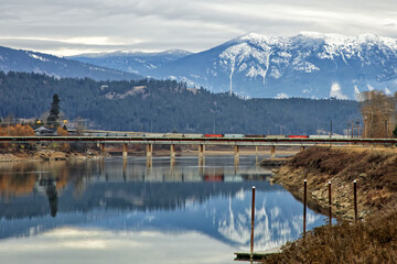Original landscaped photograph of a freight train crossing a bridge over a river with reflections and snow covered mountains
