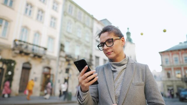 Woman with glasses wearing a coat walking down an old street and using smartphone. Slow motion