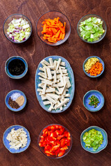 Flat lay of ready to make stir-fry ingredients: firm tofu, cut vegetables, soy sauce, spices in bowls.