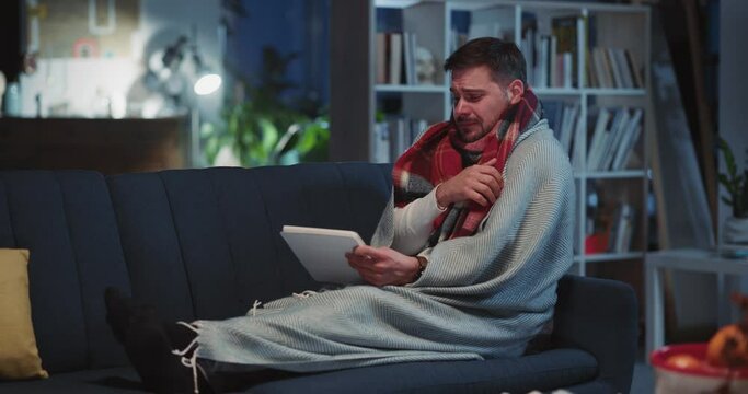 Unhealthy funny bearded man lying on sofa complaining about illness coughing and sneezing videochatting tablet computer social media at home.