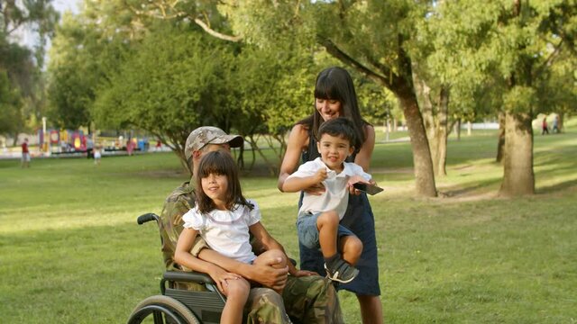 Happy mom taking selfie with two kids and disabled military dad in park, setting children on fathers lap, using cellphone. Veteran of war or leisure time with family concept