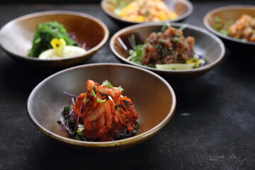 A close-up of a Korean kimchi salad on a black stony counter, composition of appetizers in a background.