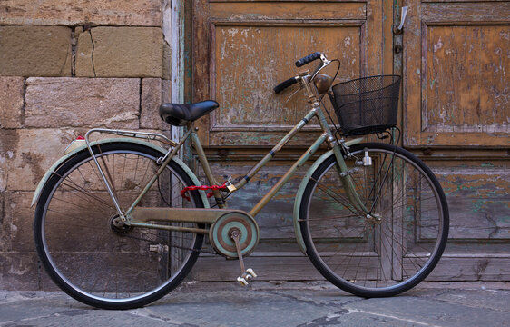 An classic, old, vintage bicycle with wicker basket rests against a weathered wall and door in Lucca, Italy
