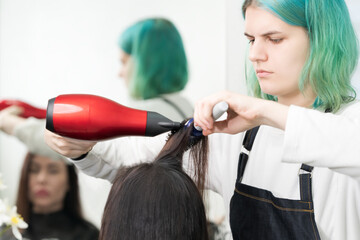 Professional hairdresser with green hair dries hair to client with red hairdryer and blue comb in beauty salon.