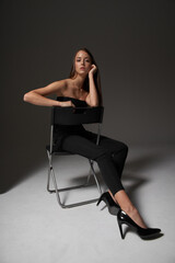 Full length studio portrait of young slim tanned caucasian girl in black jeans and bando top sitting at chair and posing against grey studio background