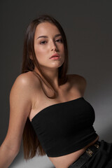 Fototapeta premium Studio portrait of young slim tanned caucasian girl in black jeans and bando top sitting and posing against grey studio background