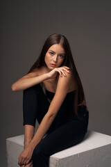 Full length studio portrait of young slim tanned caucasian girl in black jeans and bando top sitting and posing against grey studio background