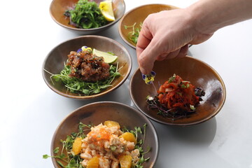 Male hand decorating a composition of appetizers: kimchi, shrimps salad, tuna, salmon tartare, seaweed salad, on white background.