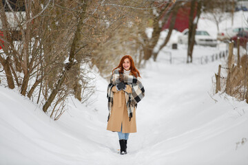 Fototapeta na wymiar Happy woman in winter snow park wearing beige coat and large plaid scarf. Romantic walk in rustic style. Female in casual classic clothes with red hairs. Redhead smiling young girl with dried flowers