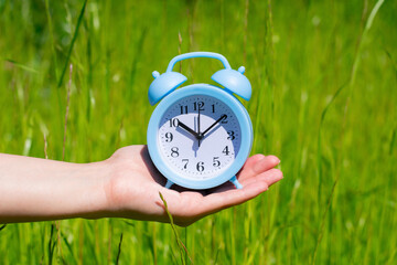 Blue alarm clock on female hand as concept of summer time coming. Green lush grass background...