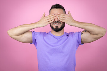 Young handsome man wearing casual t-shirt over pink background covering eyes with hands smiling cheerful and funny. Blind concept.